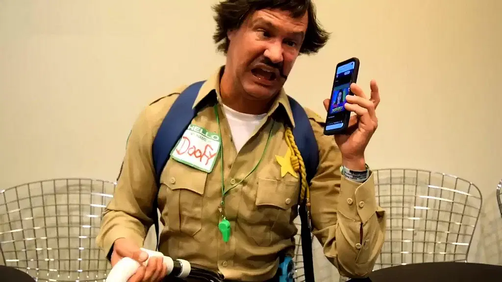 Dave Sheridan in a police uniform holding up a cell phone.